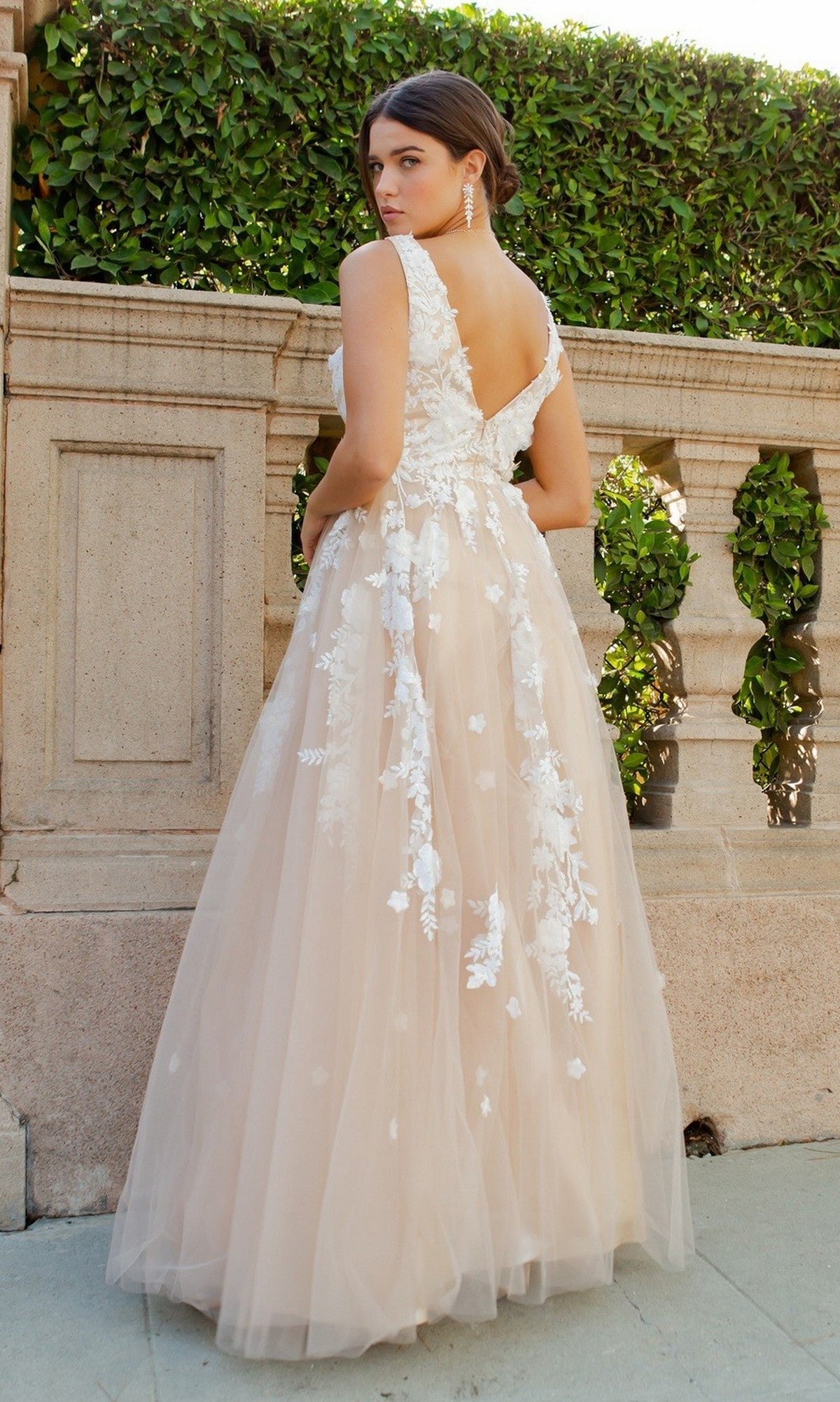 A woman in the Cinderella Couture 8040J long formal dress, featuring sleeveless styling and 3D floral lace appliques, stands outdoors by a stone railing and manicured greenery, turned slightly away from the camera.